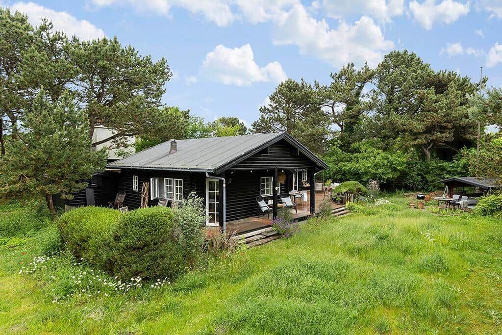 Schwarze Holzhütte mit Terrasse, umgeben von Gras und Bäumen unter blauem Himmel.