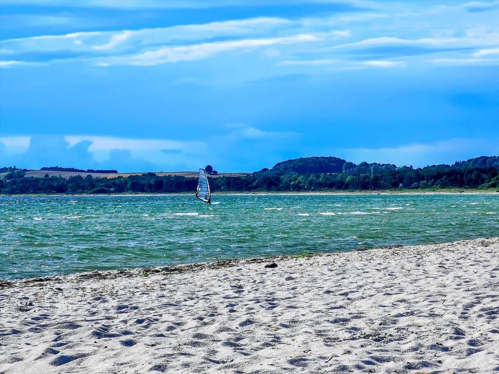 Windsurfer auf dem Wasser vor sandigem Strand und bewaldetem Ufer.