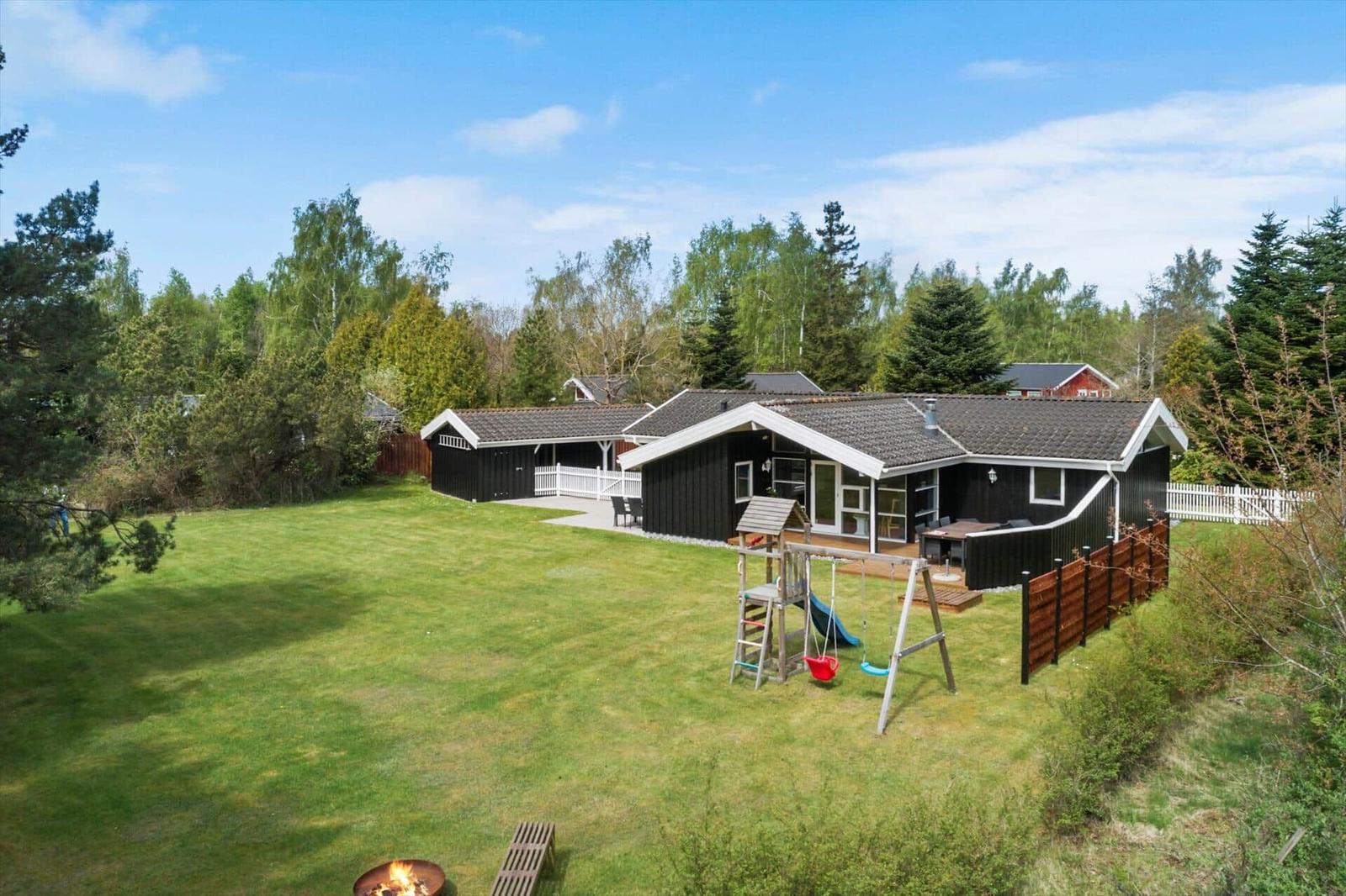 Schwarze Holzhütte mit großem Garten, Spielplatz und Grillplatz. Umgeben von Bäumen.