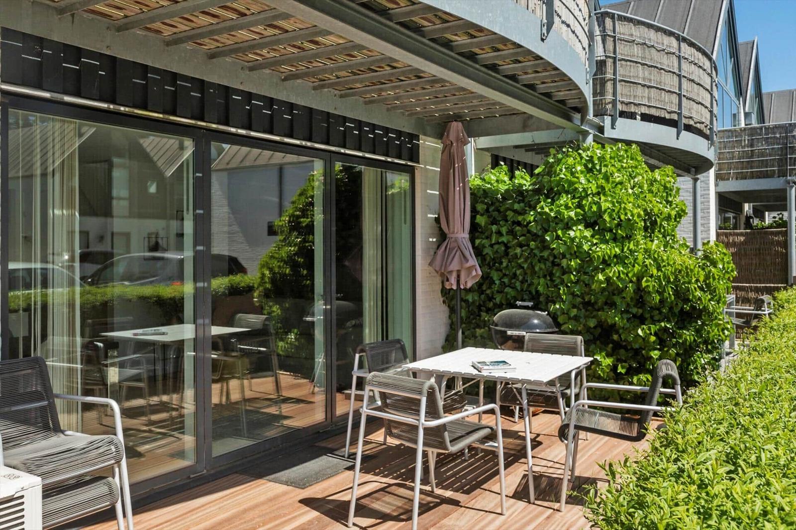 Terrasse mit Tisch, Stühlen und Grill. Glasfront und grüne Hecke sichtbar.