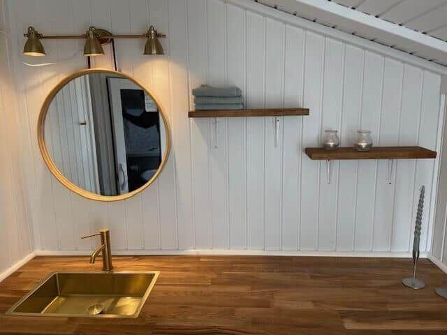 Badezimmer mit runden Spiegel, Holzregalen und goldfarbenem Waschbecken.