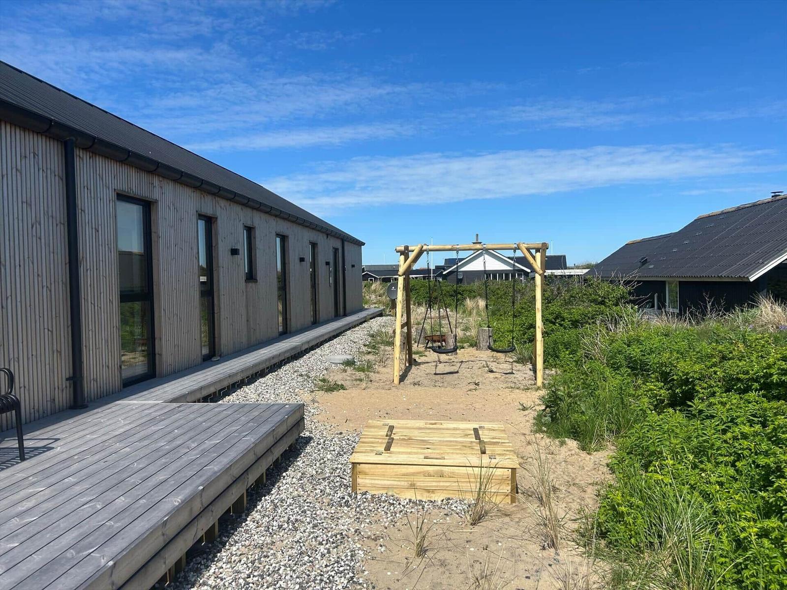 Moderne Holzhäuser mit Terrasse, Schaukel und Sandfläche im Freien.