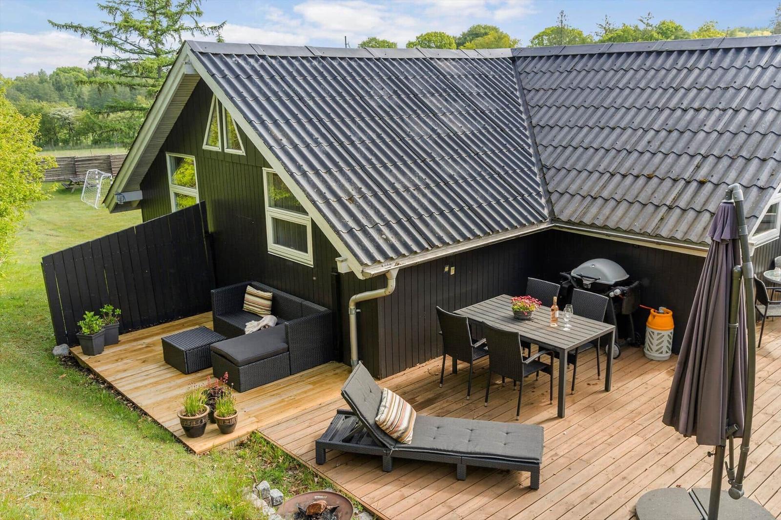 Holzterrasse mit Sitzgelegenheiten und Grillplatz vor einem Haus mit dunkler Fassade.