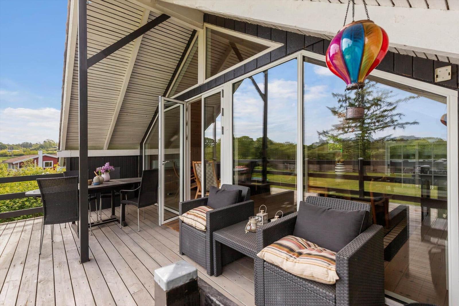 Terrasse mit Sitzgruppe, Tisch und farbenfroher Hängelampe. Glasfront öffnet zum Innenraum.