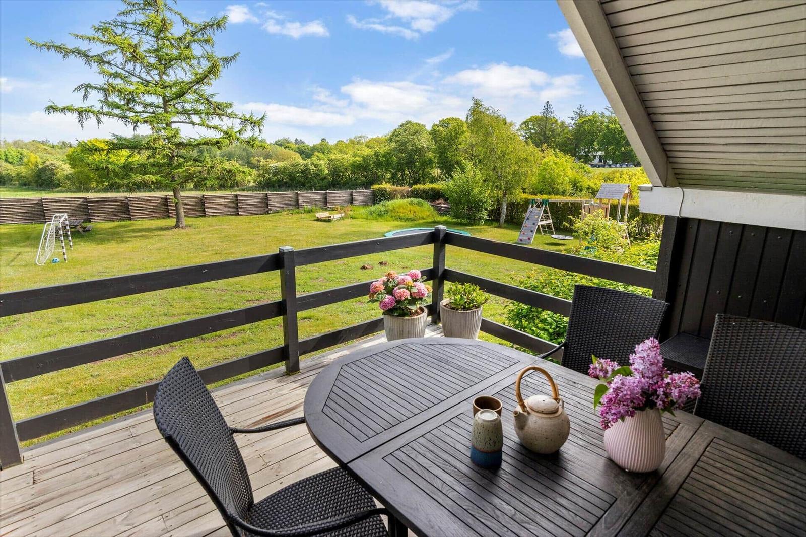 Terrasse mit Tisch und Stühlen, Blick auf grünen Garten mit Spielplatz und Bäumen.