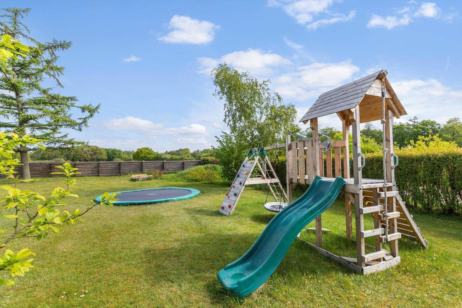 Großer Garten mit Klettergerüst, Rutsche und Sprungtrampolin. Umgeben von Bäumen und Hecke.