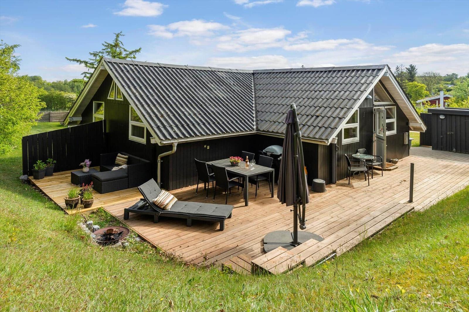 Holzterrasse mit Gartenmöbeln, Grill und Liegeplatz vor einem dunklen Holzhaus.