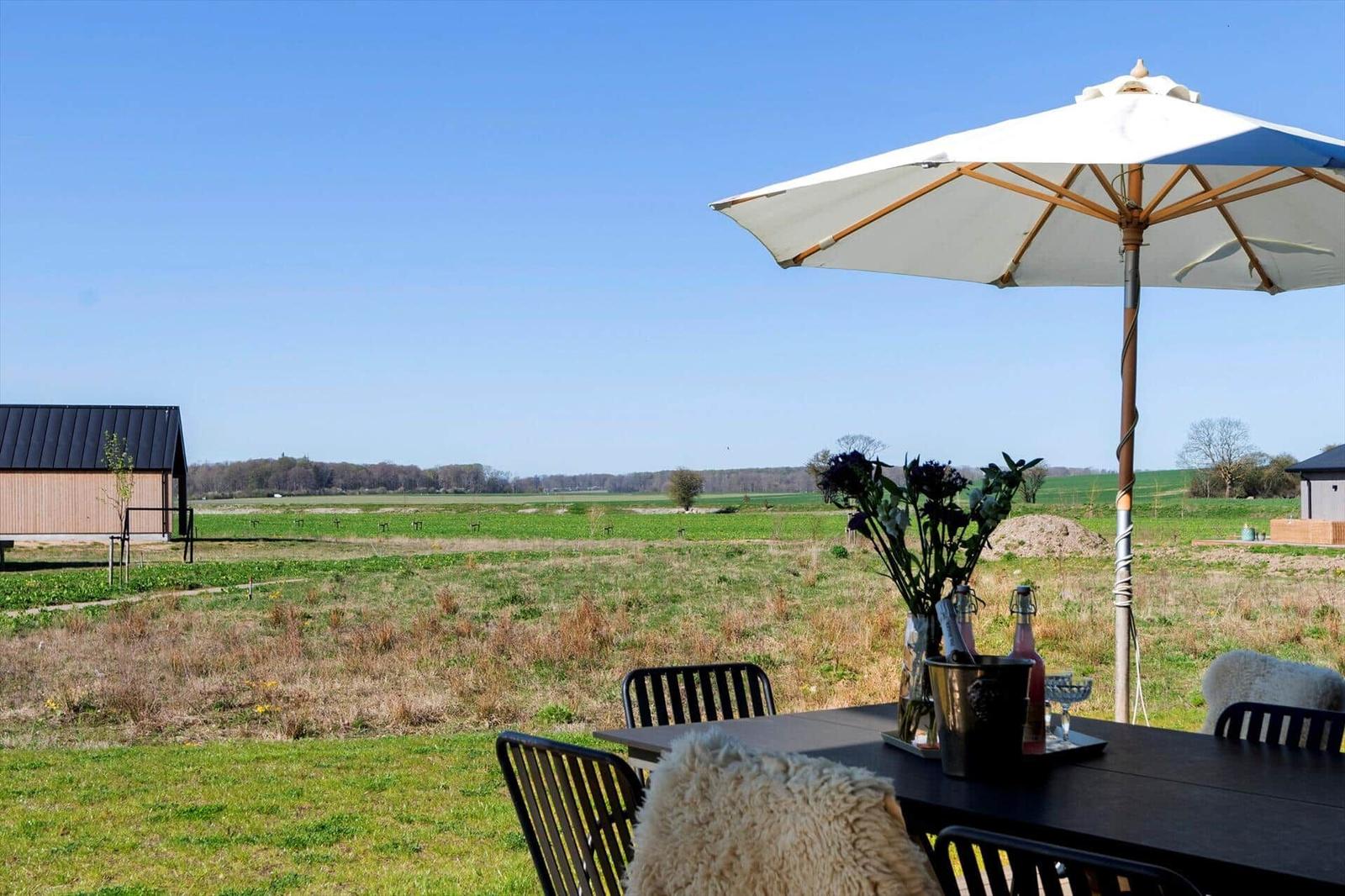 Terrasse mit Tisch, Stühlen und Sonnenschirm. Blick auf grünes Feld und Himmel.