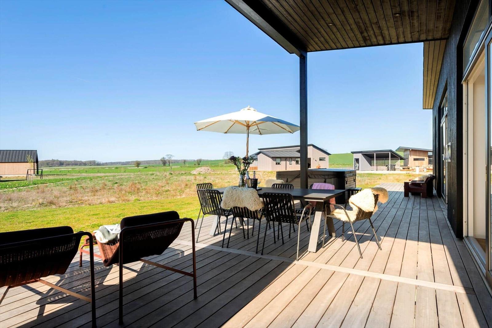 Terrasse mit Tisch, Stühlen und Sonnenschirm. Blick auf grüne Wiesen und Gebäude.
