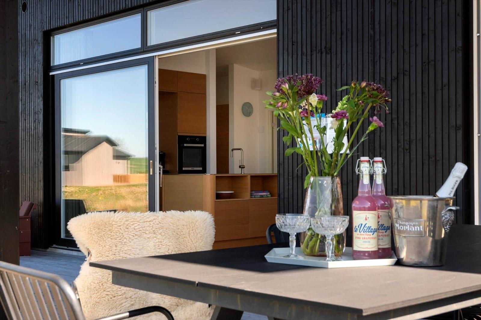 Terrasse mit Tisch, Stühlen und Blumen. Durchgehendes Fenster zeigt Küchenbereich.