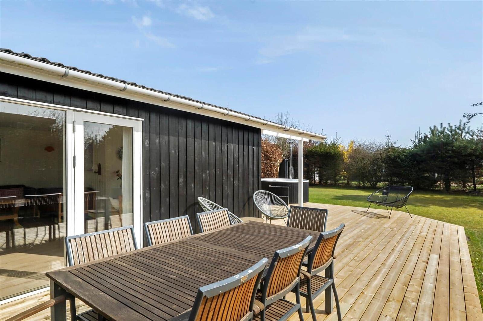 Deck with wooden table and chairs, adjacent garden and holiday home with glass doors.