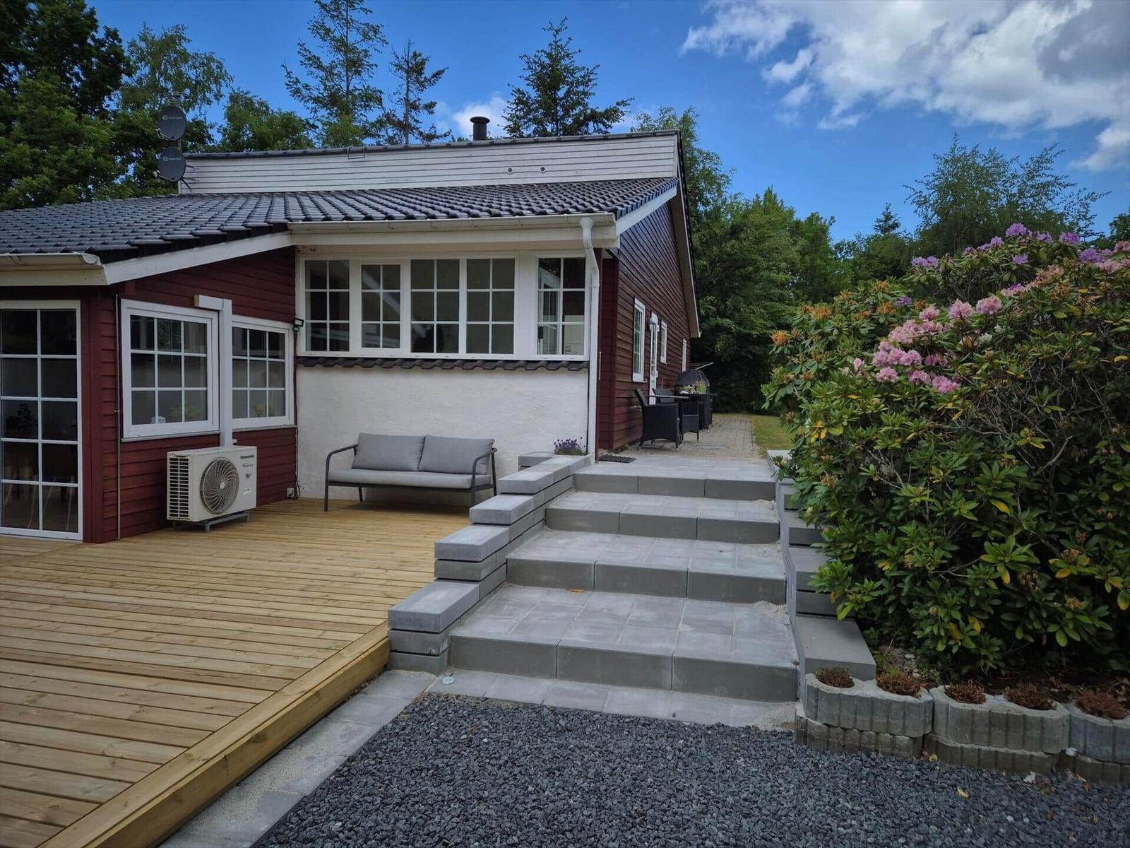 House with terrace, bench, and steps to entrance. Surrounded by greenery and flowers.