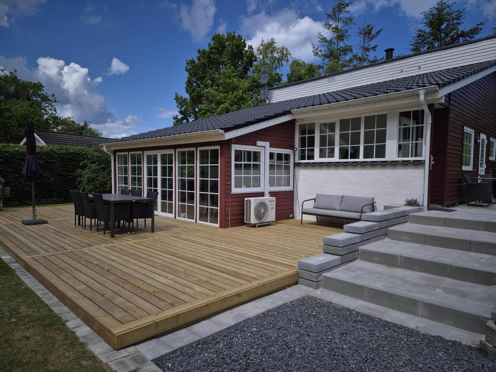 Deck with table, chairs, and sofa in front of a house with red wood siding and white windows.