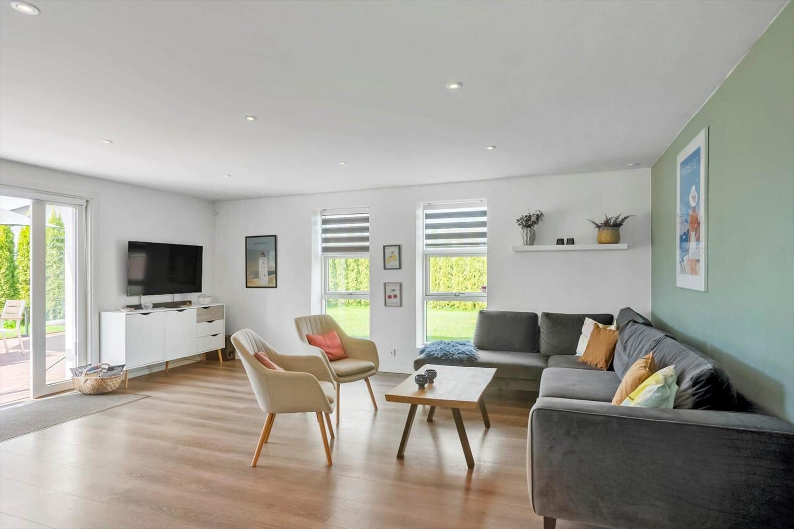 Wohnzimmer mit Sofa, zwei Sesseln, Couchtisch und Fernseher. Holzboden und große Fenster.