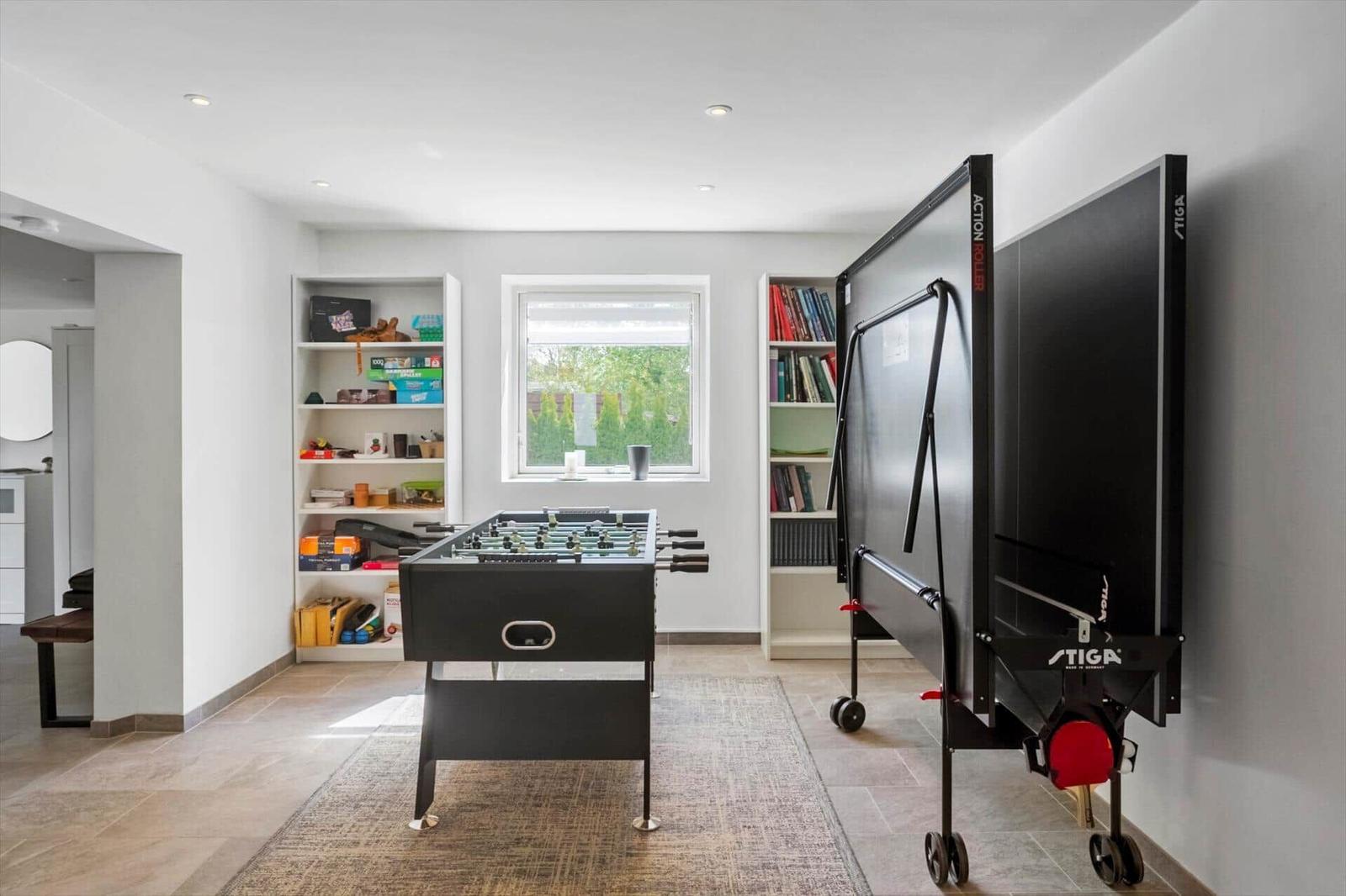 Raum mit Tischfußball und aufstellbarem Tischtennis-Tisch. Bücherregale und Fenster mit Blick auf Garten.