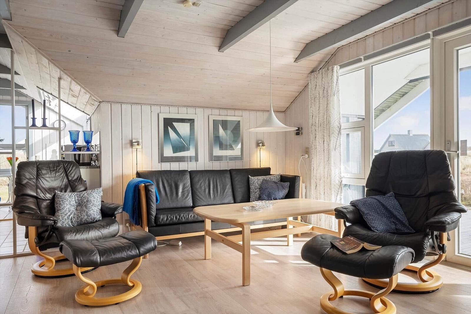 Wohnzimmer mit schwarzen Ledersofas und zwei Sessel. Holztisch und große Fenster.