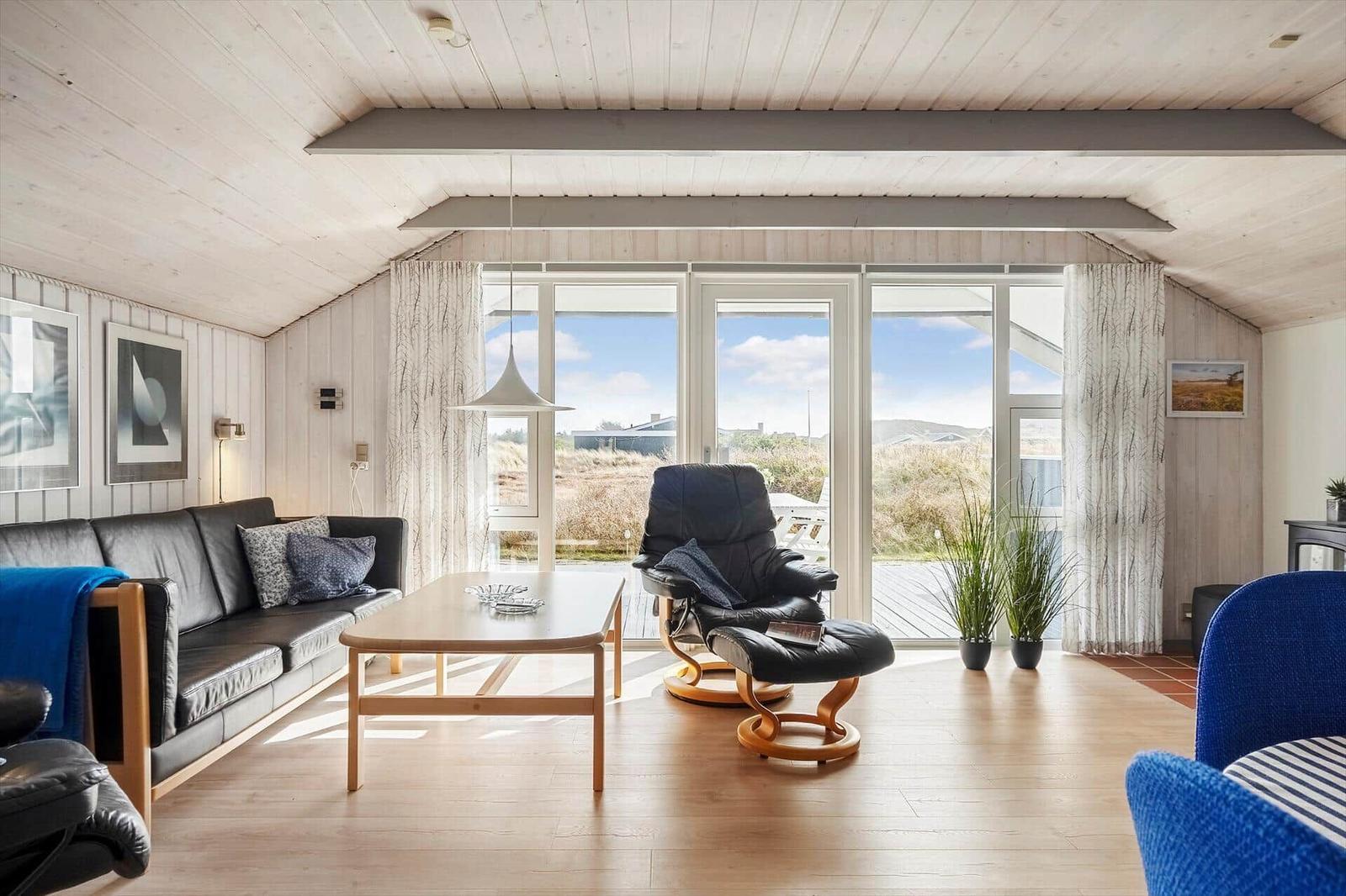 Wohnzimmer mit Sofa, Sessel und Tisch. Große Fenster bieten Blick auf Außengelände.