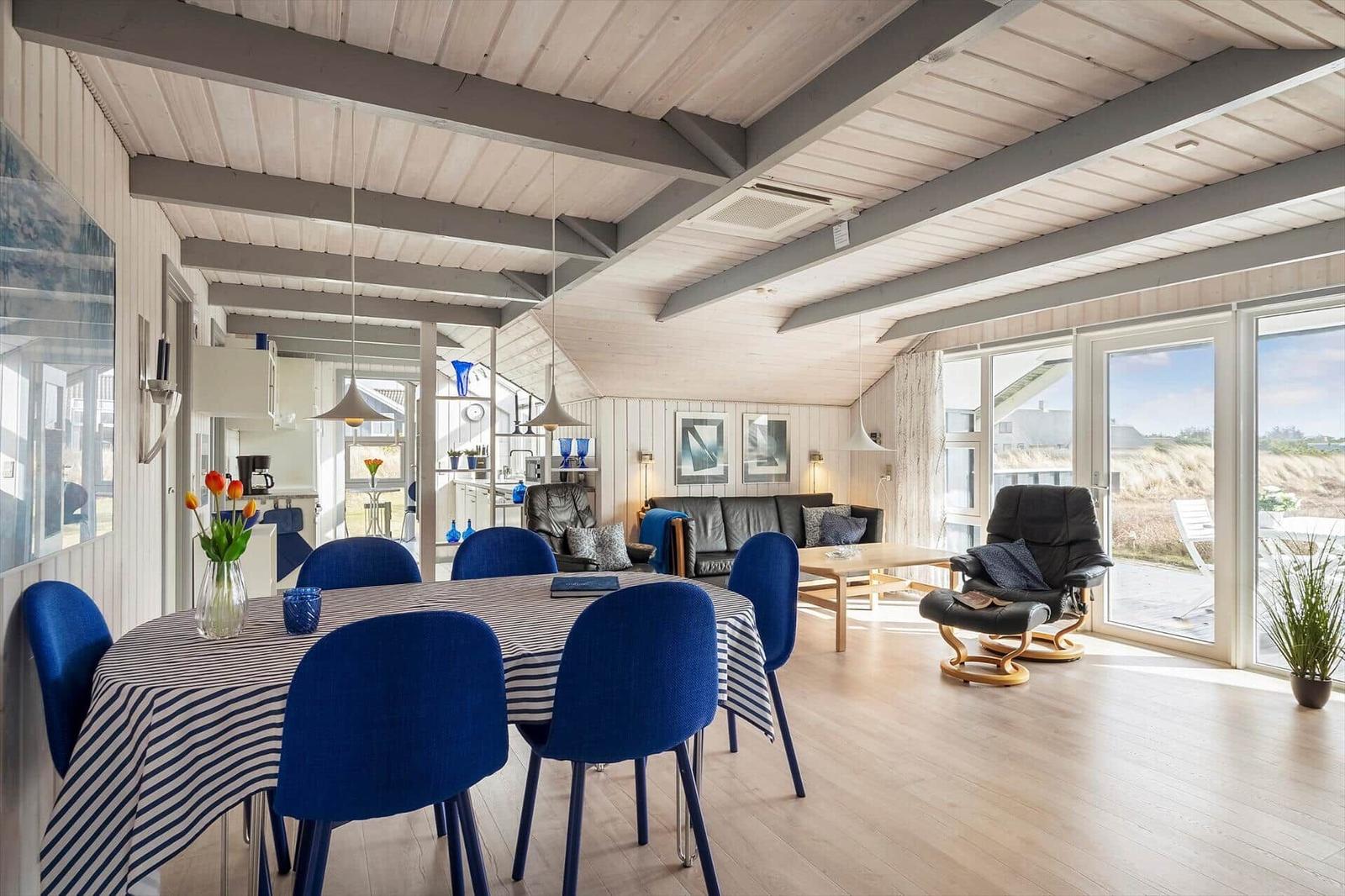 Essbereich mit blauen Stühlen und Tischdecke. Wohnbereich mit Sofa und Sessel. Holzboden und große Fenster.
