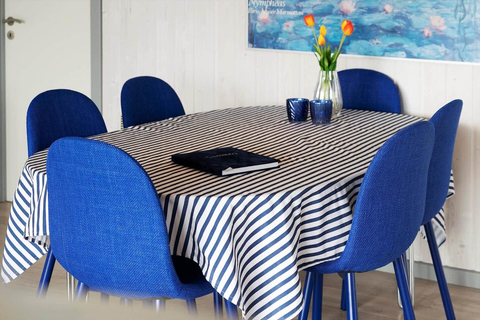 Runder Tisch mit gestreifter Tischdecke und blauen Stühlen. Hintergrund: Wand mit Bild und Tulpen.