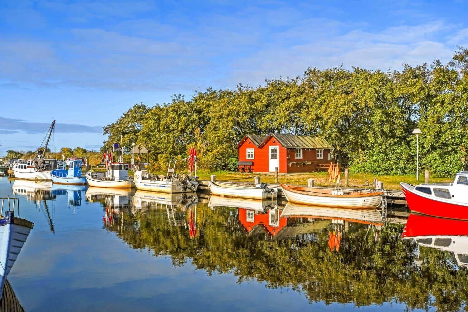 Roter Holzschuppen am Ufer mit Booten und Spiegelung im Wasser.
