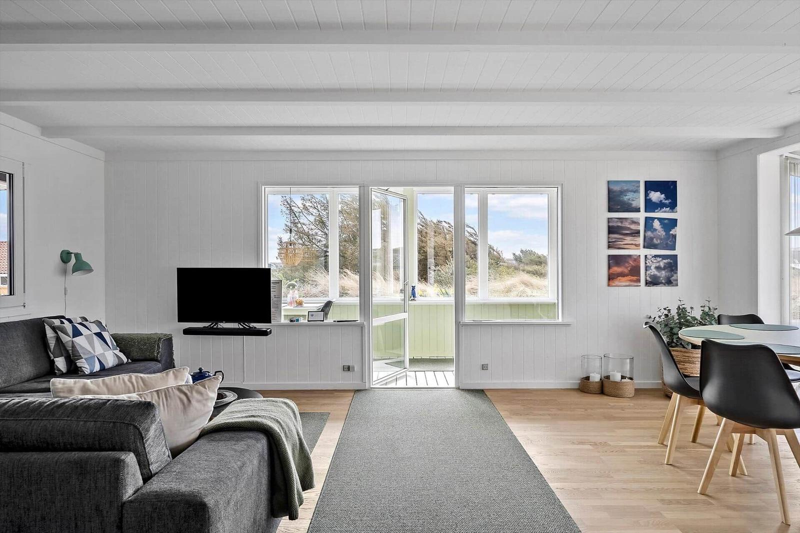 Wohn- und Essbereich mit Sofa, TV, Tisch und Stühlen. Durchgang zur Terrasse mit Blick auf Grasland.
