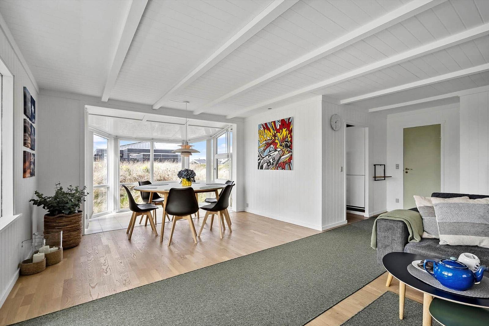 Wohn- und Essbereich mit Holzboden, Sofa, Tisch und Stühlen. Große Fensterfront mit Blick nach draußen.