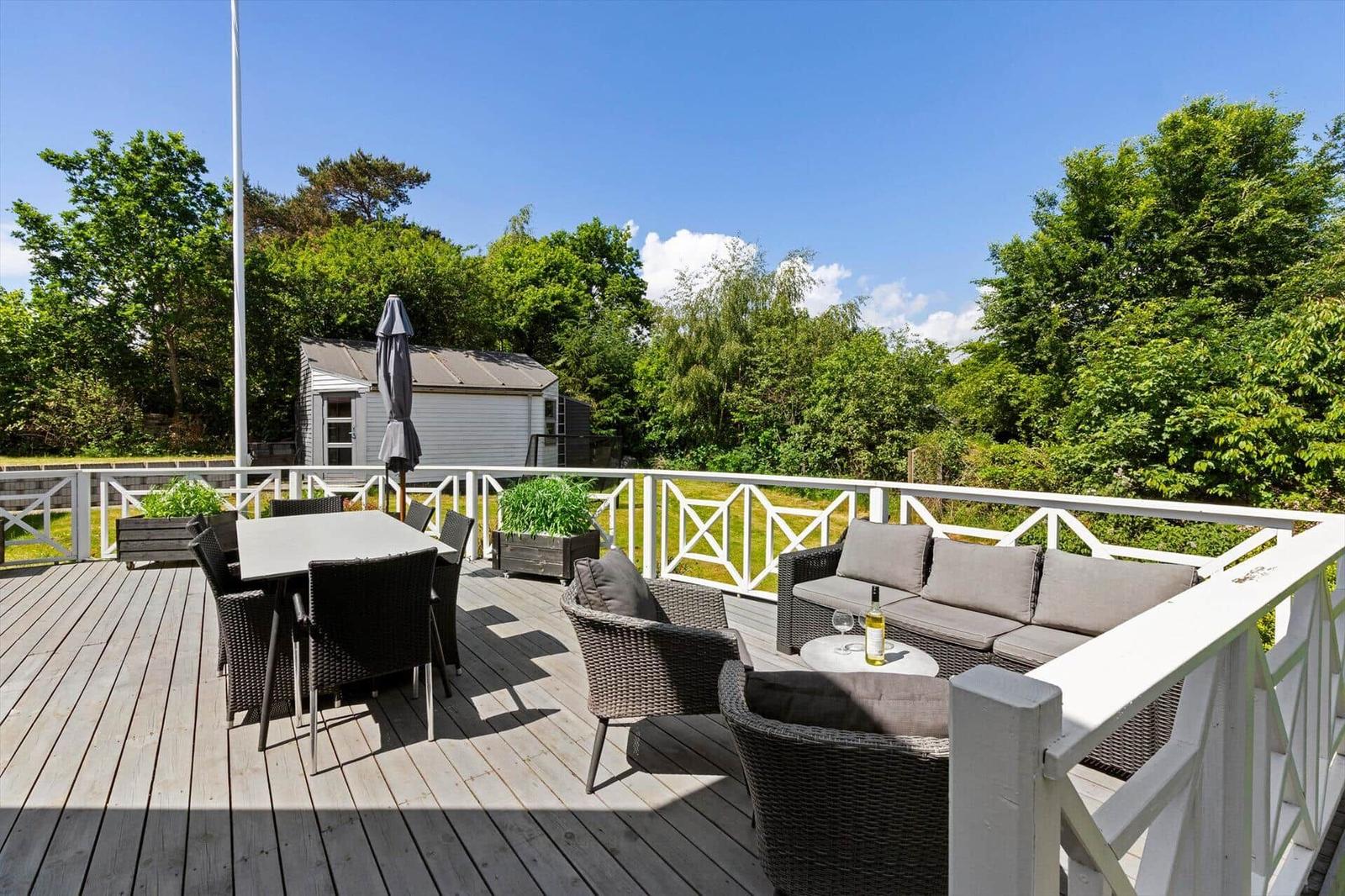 Deck with seating, table, and chairs. Background: green areas and small house.