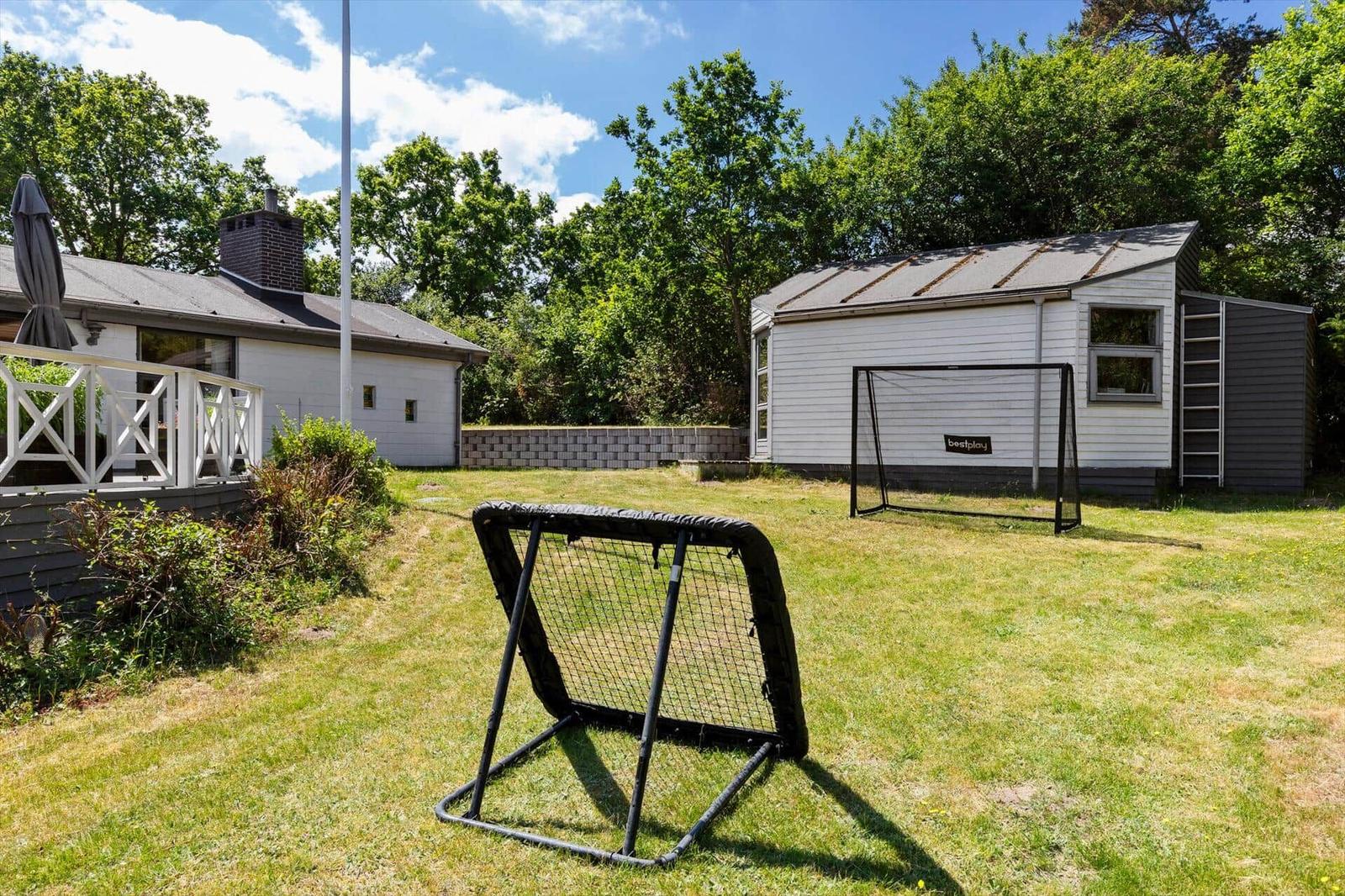Backyard with soccer goal and small house. Grass area and trees in the background.