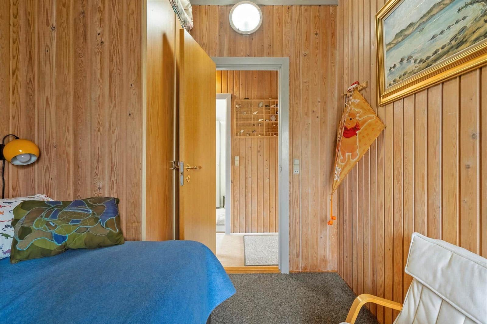Schlafzimmer mit Holzwand, Bett, Stuhl und Wandbild. Tür führt in einen Flur.