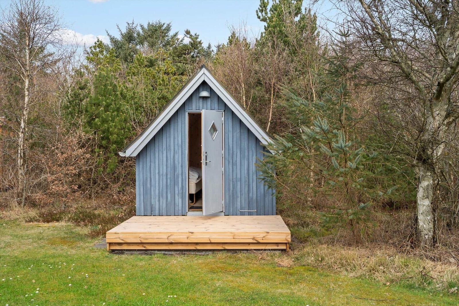 Ein kleines blaues Holzhaus mit offener Tür auf einer Holzterrasse im Wald.
