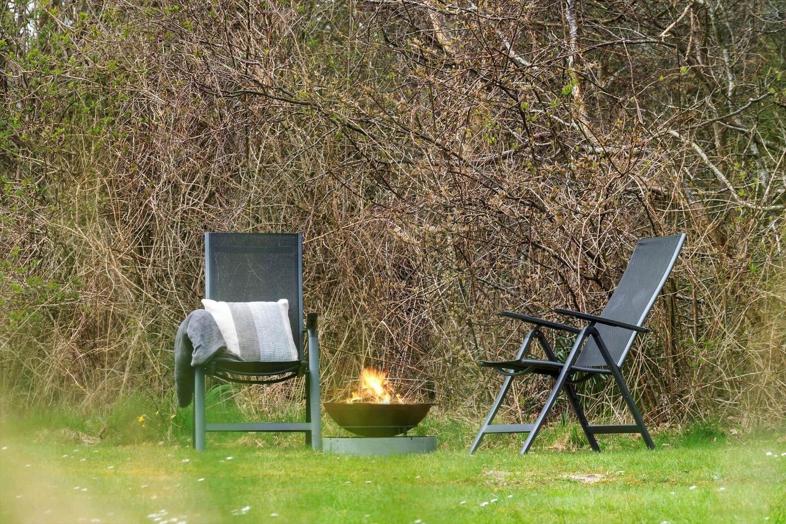 Zwei Gartenstühle und ein Feuerkorb stehen auf einer Wiese vor Sträuchern.