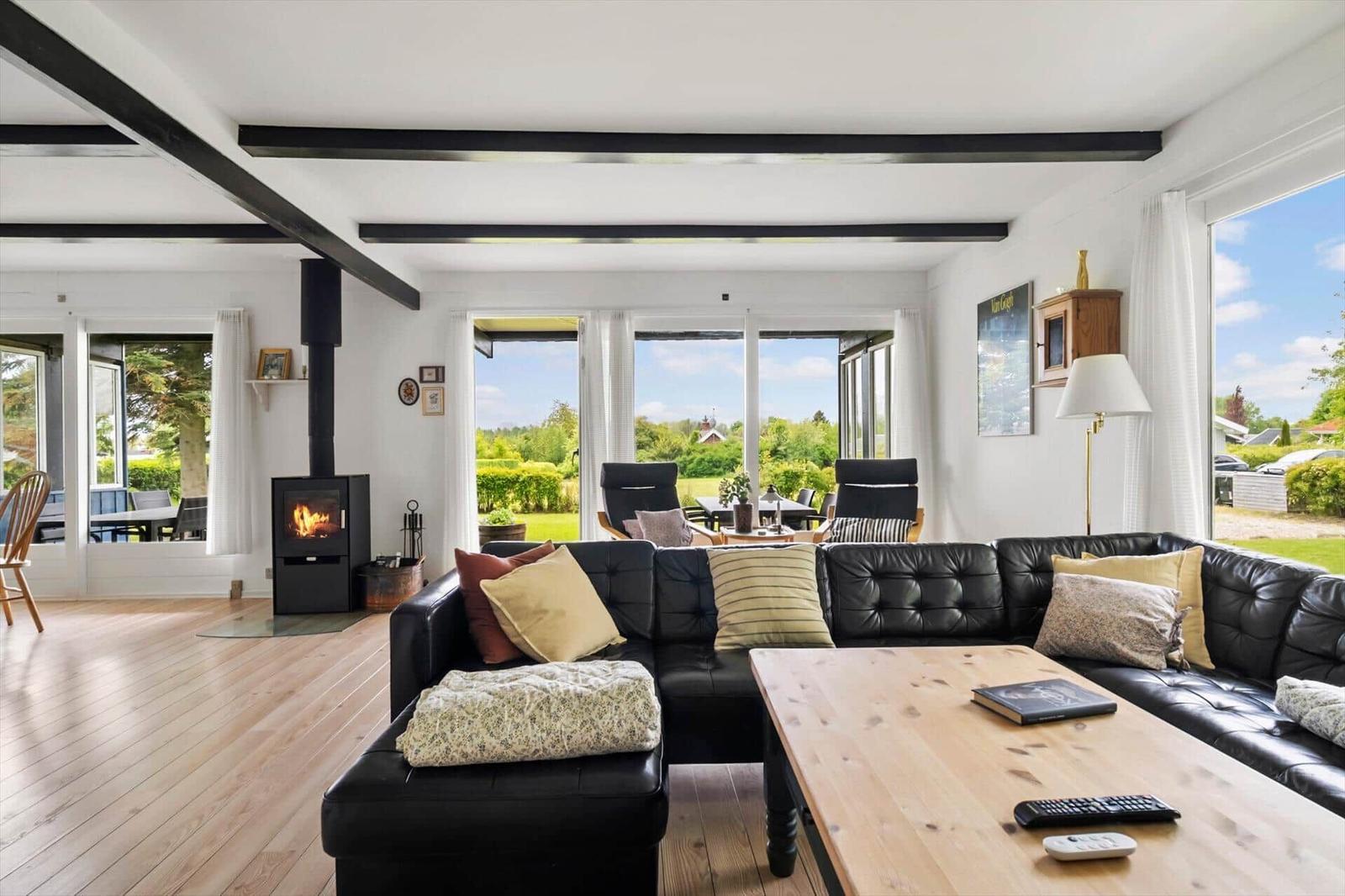 Living room with black leather sofa, wooden table, and fireplace. Large windows overlook the garden.
