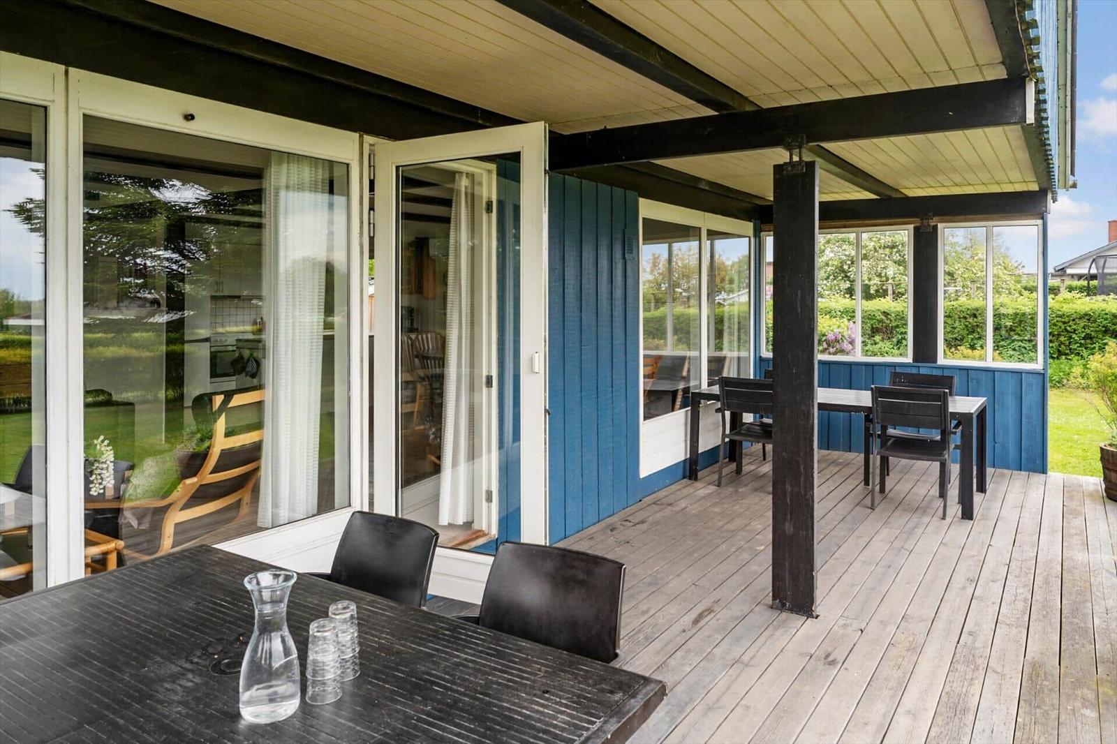 Deck with table and chairs, glass walls and blue siding, view of garden.