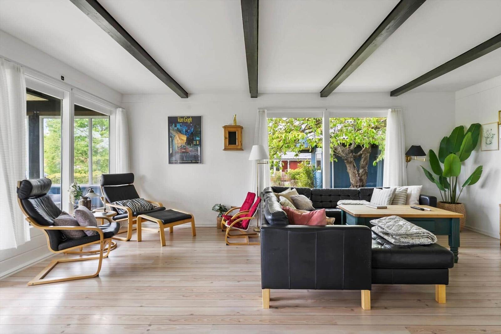 Living room with black leather furniture, wooden floor, and large windows. Beams and plants are visible.