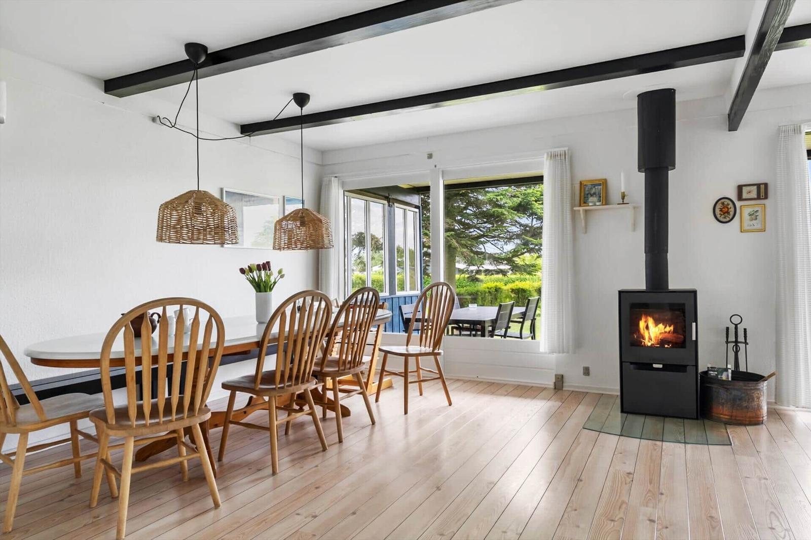 Dining room with wooden table, chairs, and wood stove. Large windows overlooking the garden.