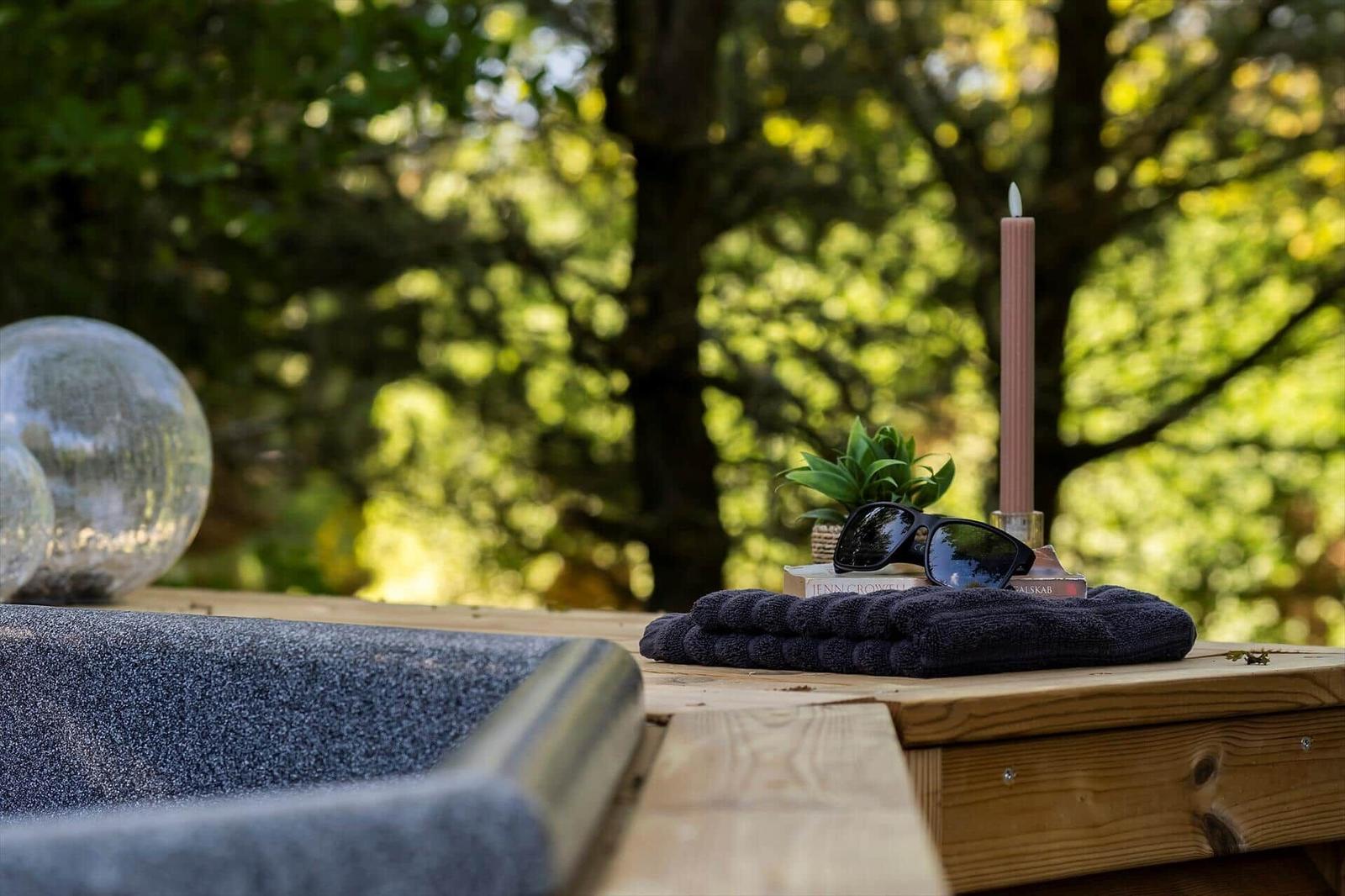 Holztisch mit dunkler Handtuch, Sonnenbrille, Kerze und Pflanze vor Waldhintergrund.