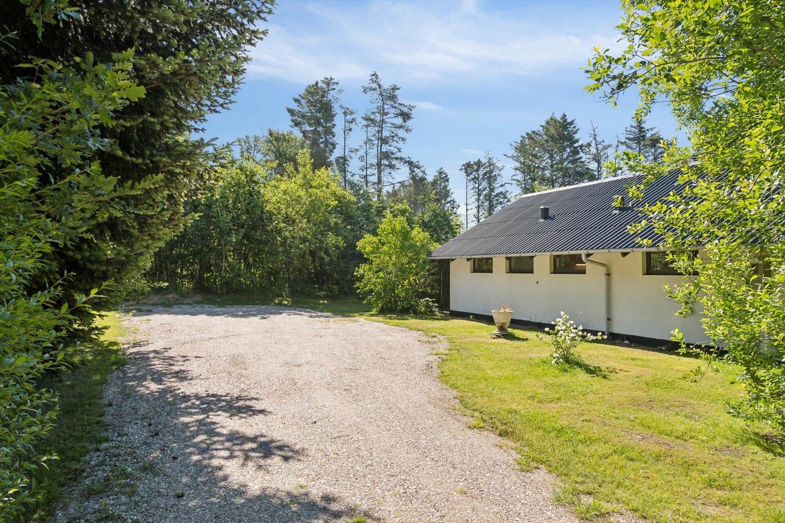 Haus mit dunklem Dach und weißen Wänden steht in grüner Umgebung mit Wald und Kiesweg.