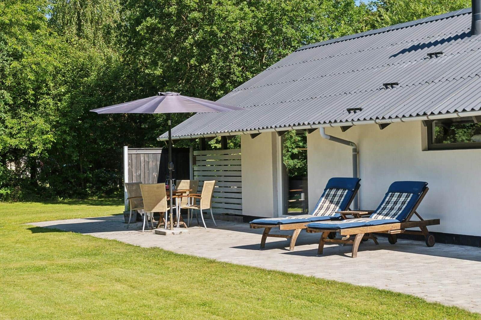 Terrasse mit Sonnenliegen, Tisch und Stühlen unter einem Sonnenschirm. Hintergrund: grüner Garten und Gebäude.