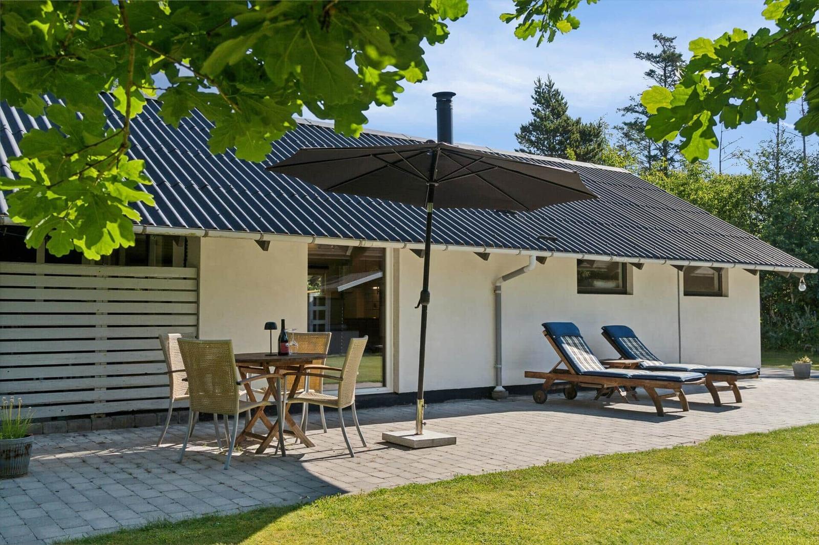 Außenbereich mit Tisch, Stühlen, Sonnenschirm und Liegestühlen vor einem Haus.