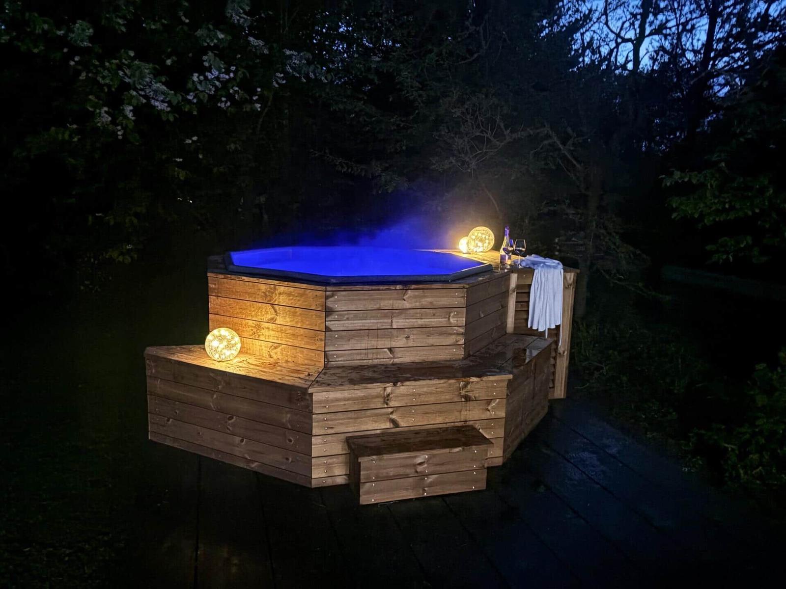 Ein hölzerner Whirlpool mit blauer Beleuchtung und Dampf, umgeben von Bäumen und Lichtkugeln.