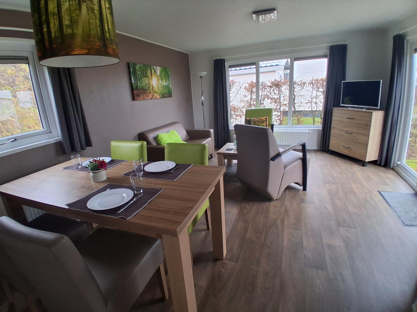 Wohn- und Essbereich mit Tisch, Stühlen, Sofa und TV. Holzdielen und große Fenster.