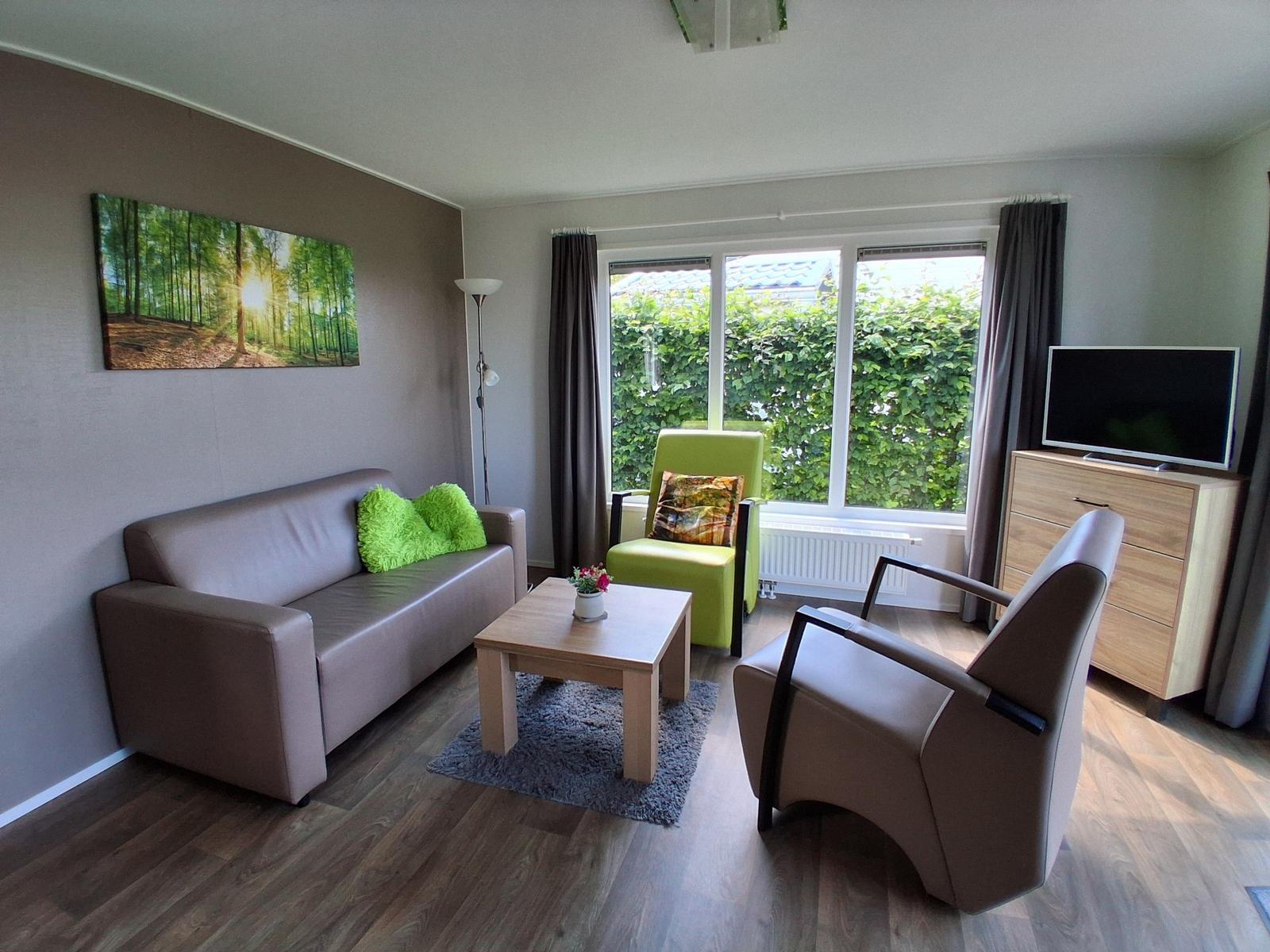 Wohnzimmer mit Sofa, Sessel und TV. Große Fenster mit Blick auf grüne Hecke.