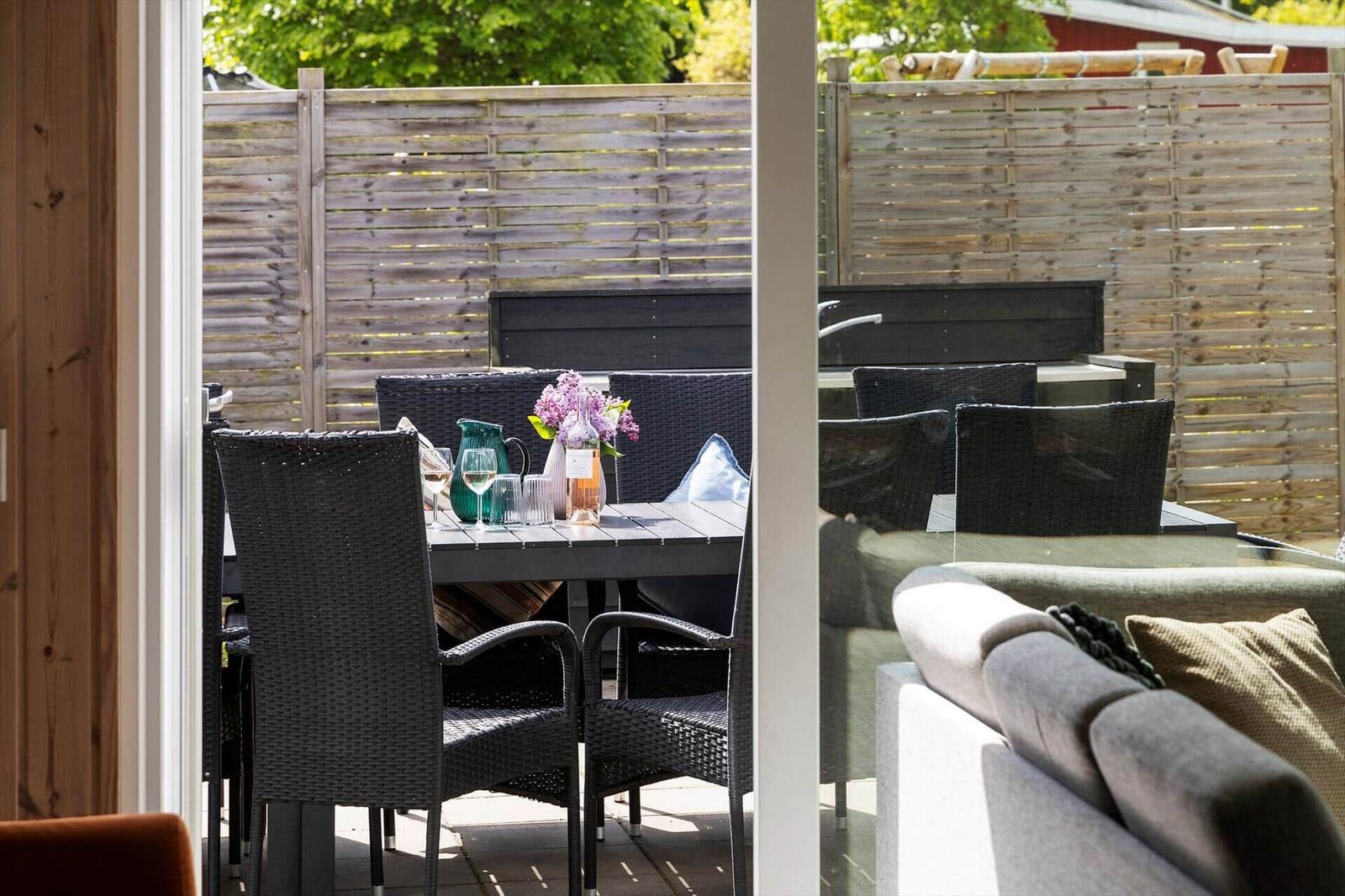 Terrasse mit Tisch, Stühlen und Grill. Hintergrund: Holzzaun und grüne Bäume.
