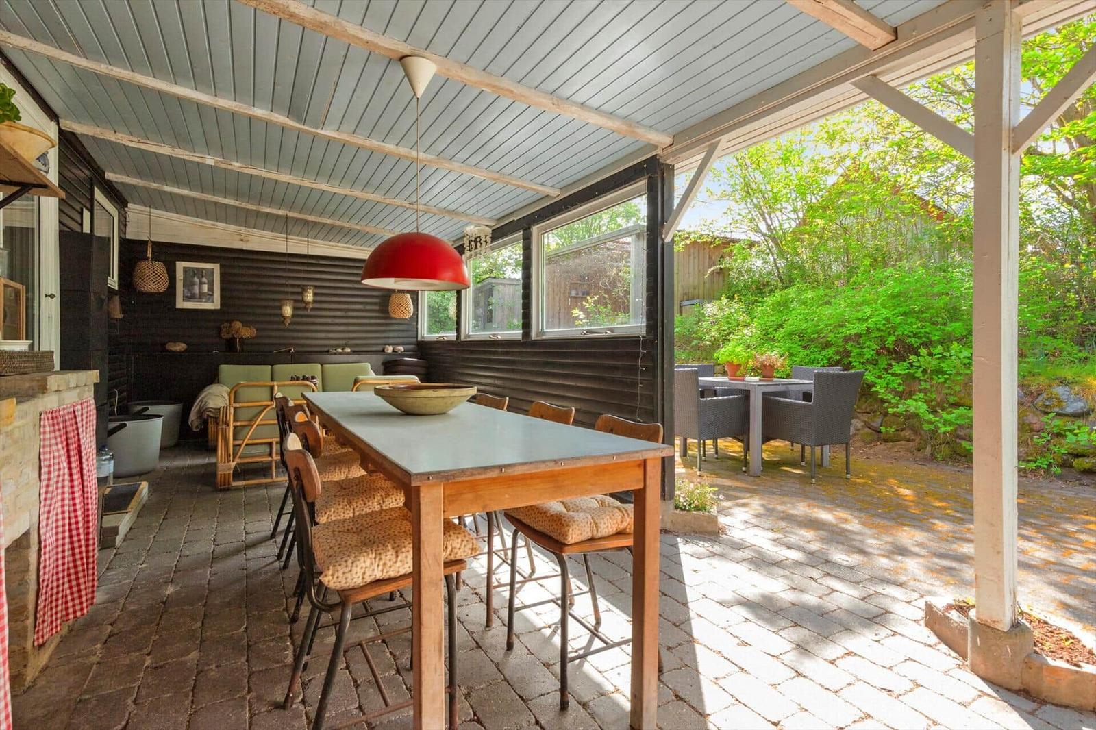 Terrasse mit Tisch, Stühlen und Blick auf grünen Garten. Decke mit Holzbalken und roter Lampe.