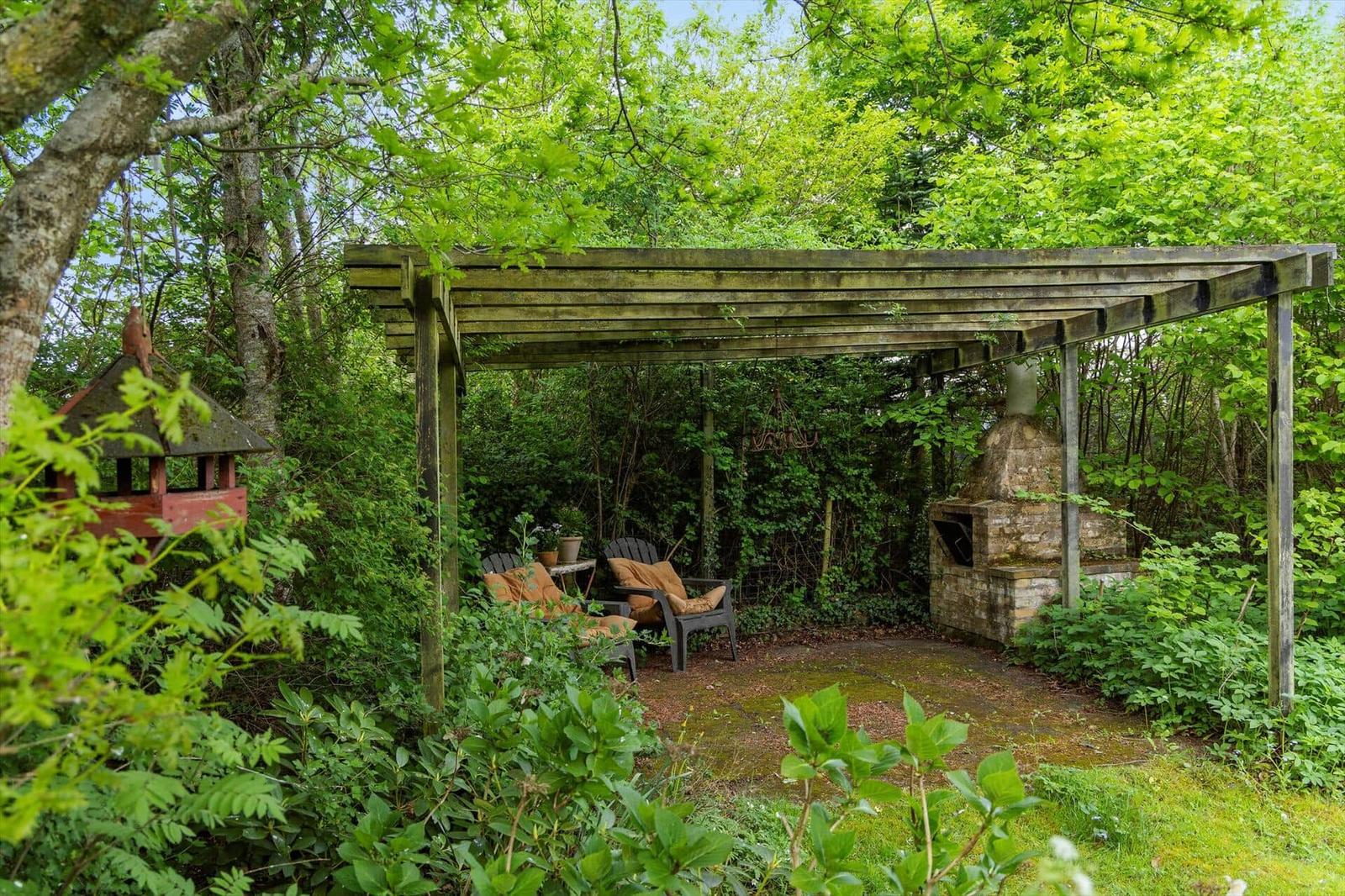 Gartenbereich mit Holzüberdachung, Sitzgelegenheiten und Steinofen zwischen Bäumen.
