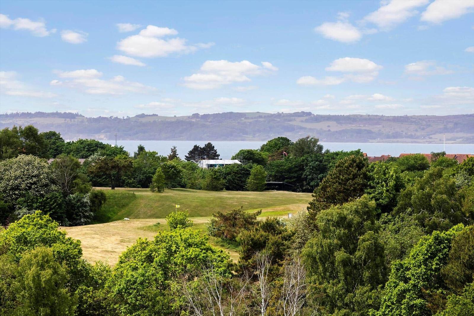 Weitläufiger Golfplatz mit Blick auf Wasser und Hügel unter blauem Himmel.