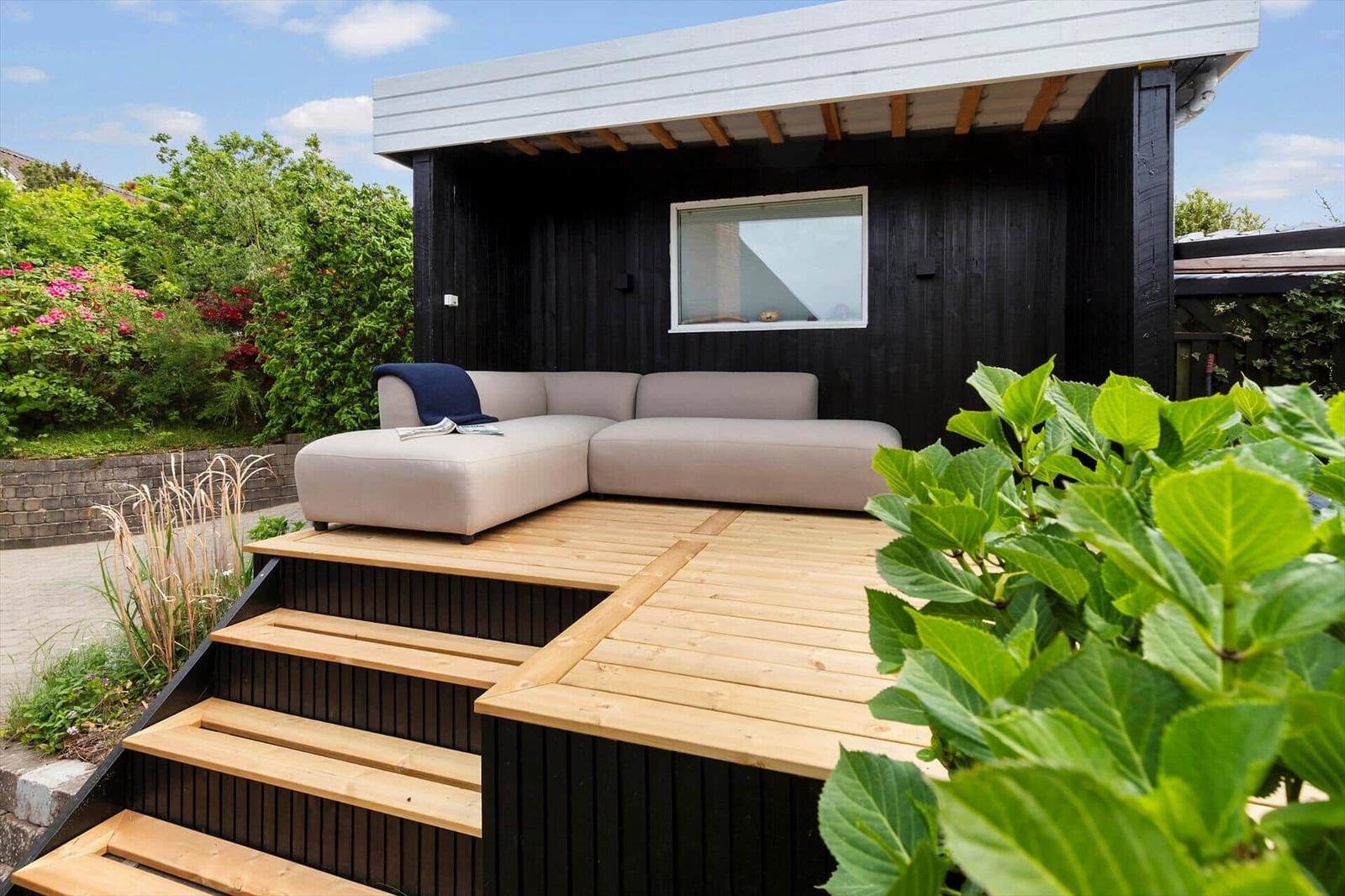 Terrasse mit grauem Sofa und Holzstufen vor schwarzem Holzhaus mit Fenster.