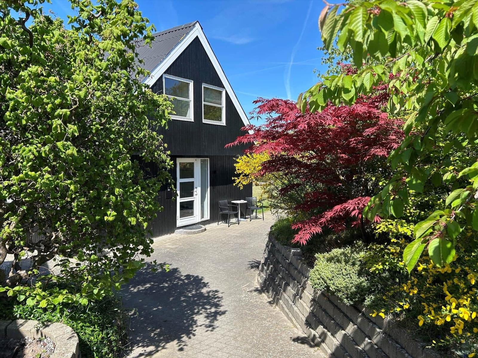 Schwarzes Haus mit weißer Tür und Gartenweg. Rote und grüne Bäume umgeben den Eingang.