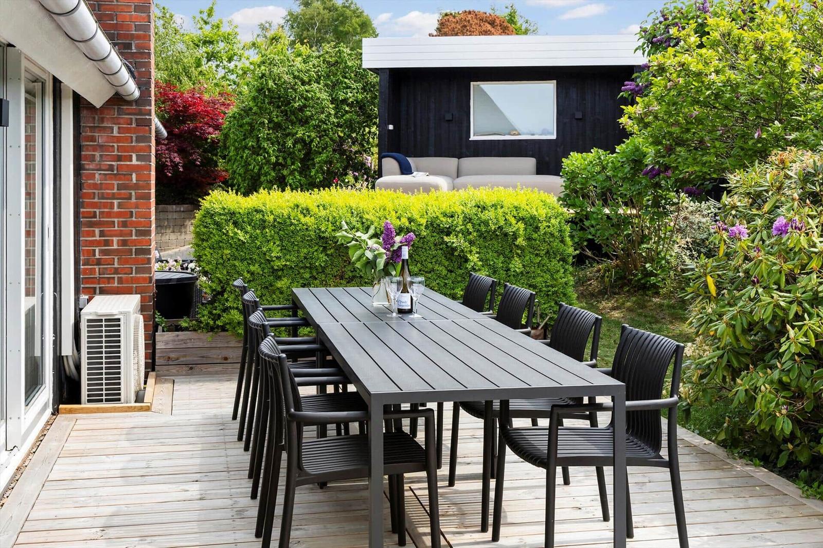 Terrasse mit langem Tisch und Stühlen, umgeben von grünen Sträuchern und einem Gartenhaus.