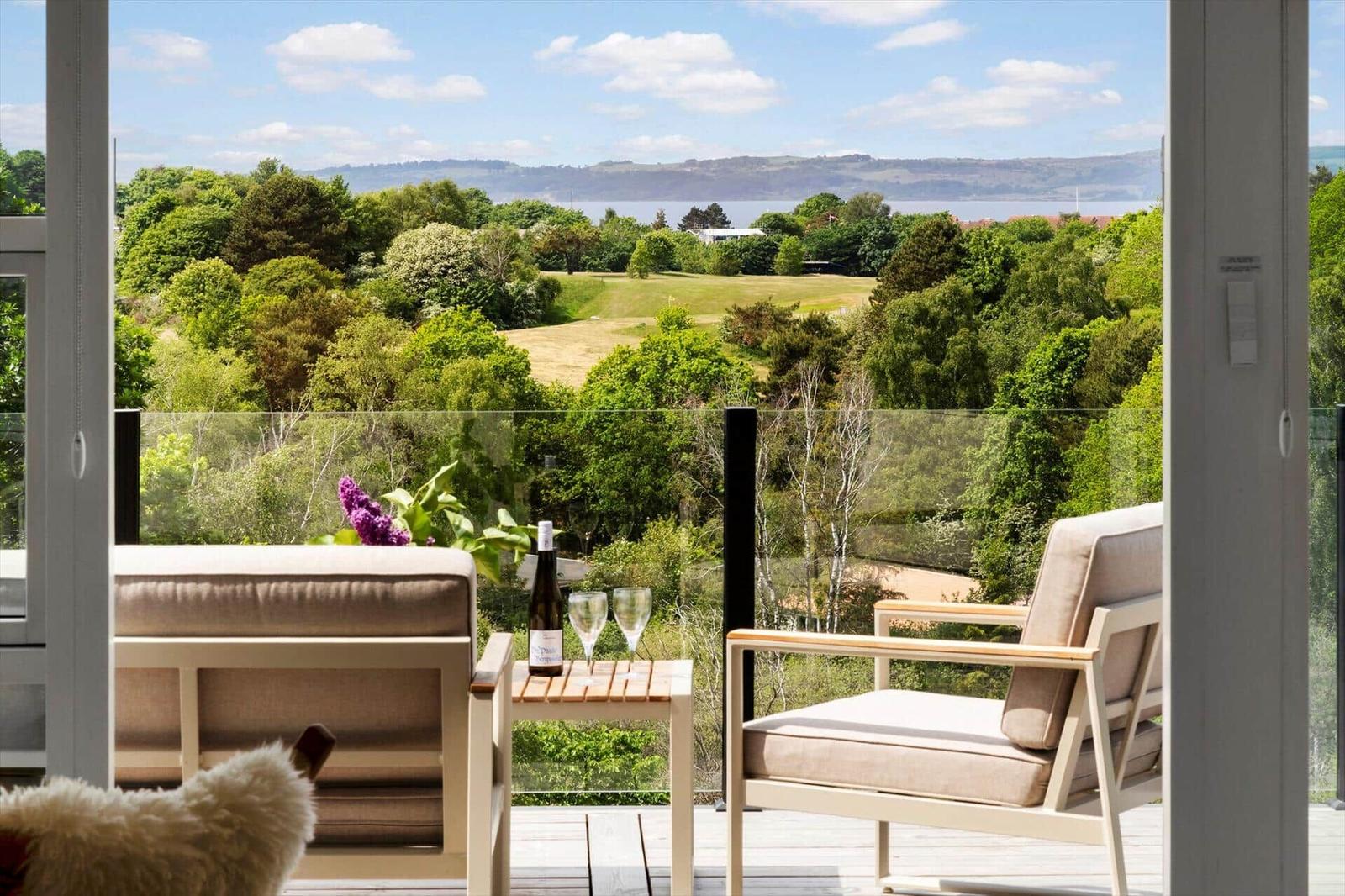 Balcony with two chairs and table, overlooking greenery and water.